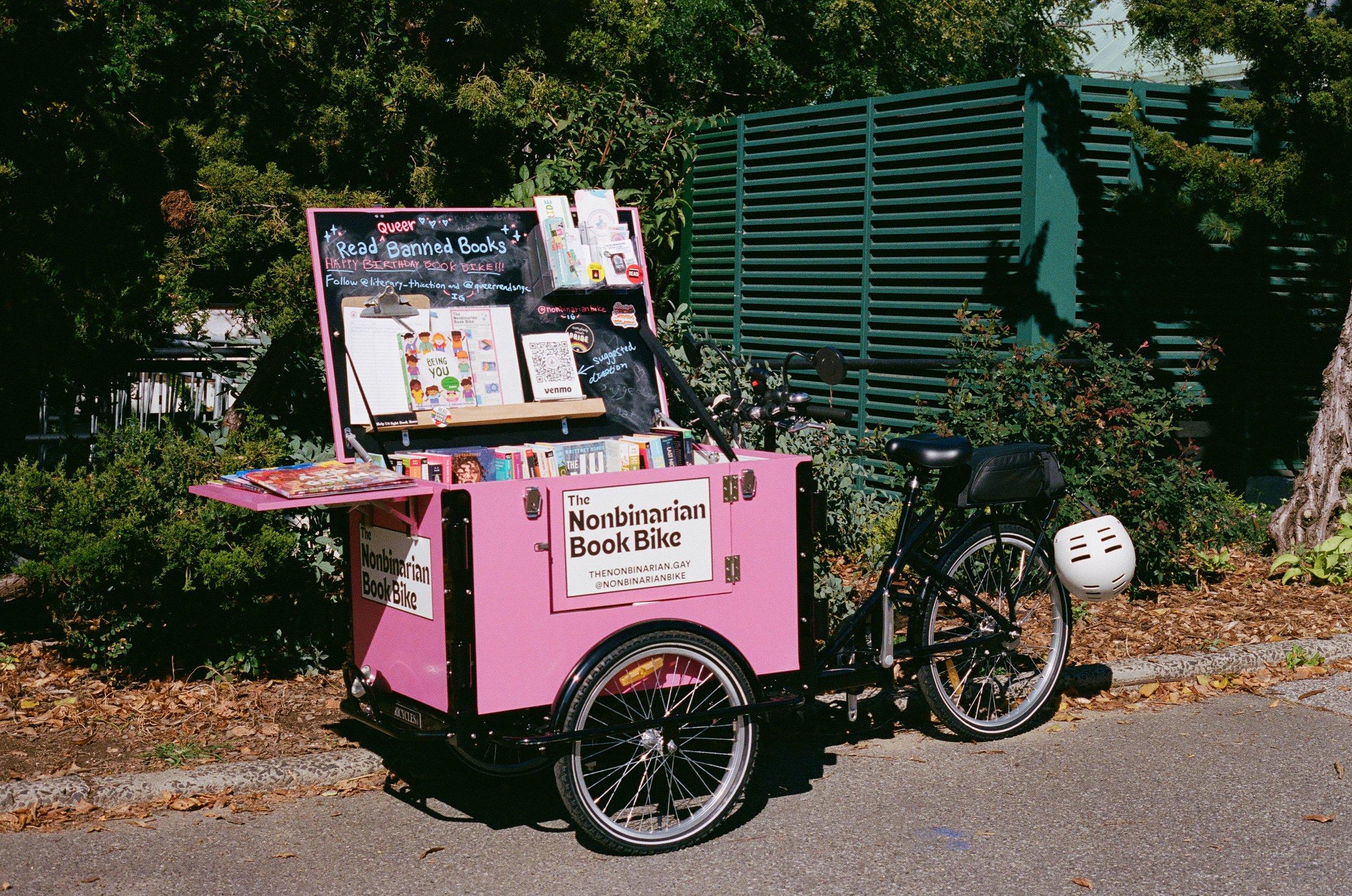 The Nonbinarian Book Bike is a bright pink trailer attached to the front of a bicycle which provides queer banned books to the NYC community.